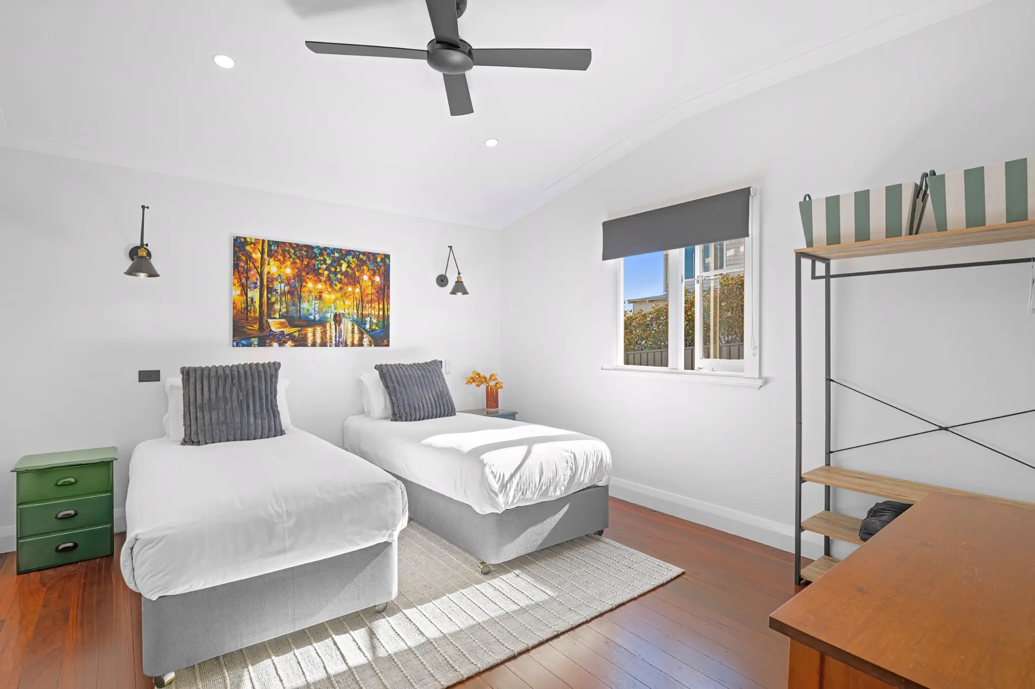 Second bedroom with two long single beds, ceiling fan, colourful artwork and study desk on jarrah floors