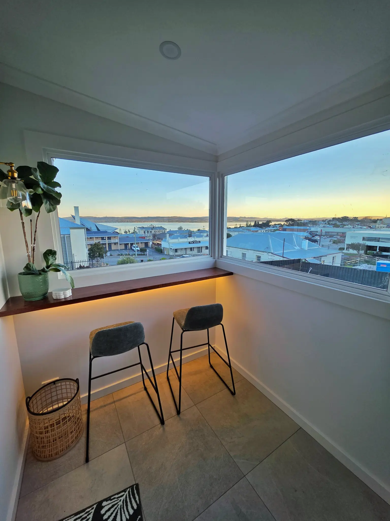 View bar at dusk with two stools and warm under-bench glow, harbour and city at sunset through corner windows