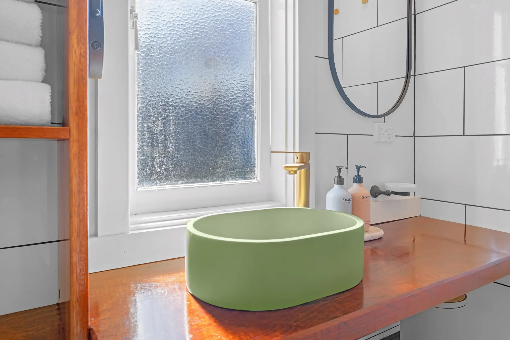 Sage green vessel basin on jarrah vanity with gold tap, round mirror and white subway tiles