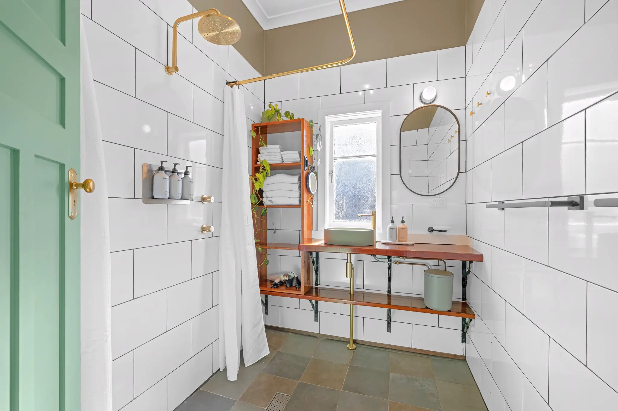 White subway-tiled bathroom with gold overhead shower, timber shelf with towels and green door