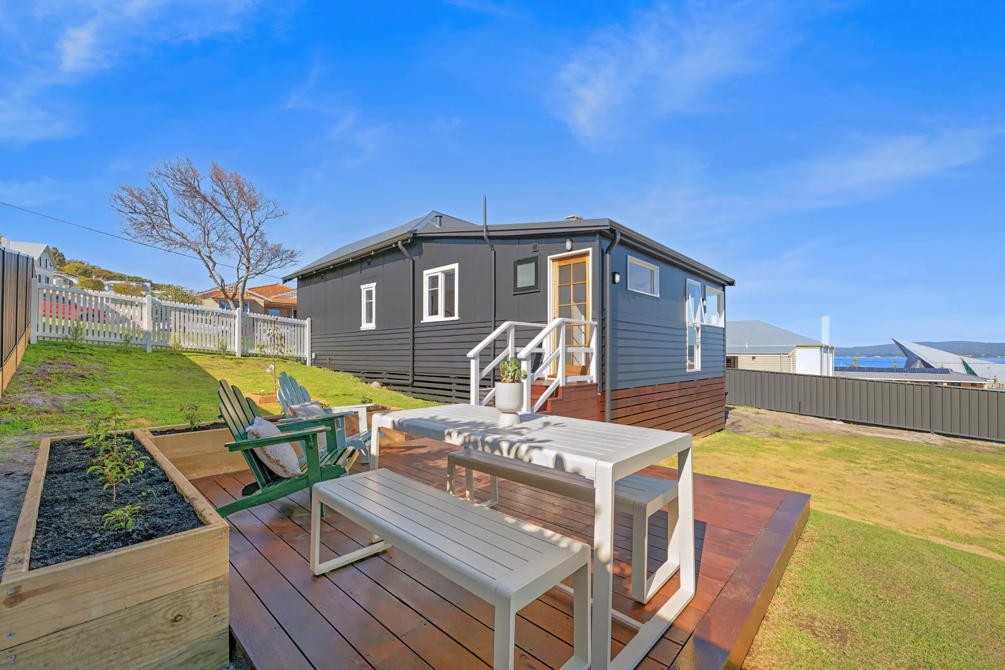 Rear of dark cottage from backyard showing timber deck, raised garden beds, lawn and harbour glimpse