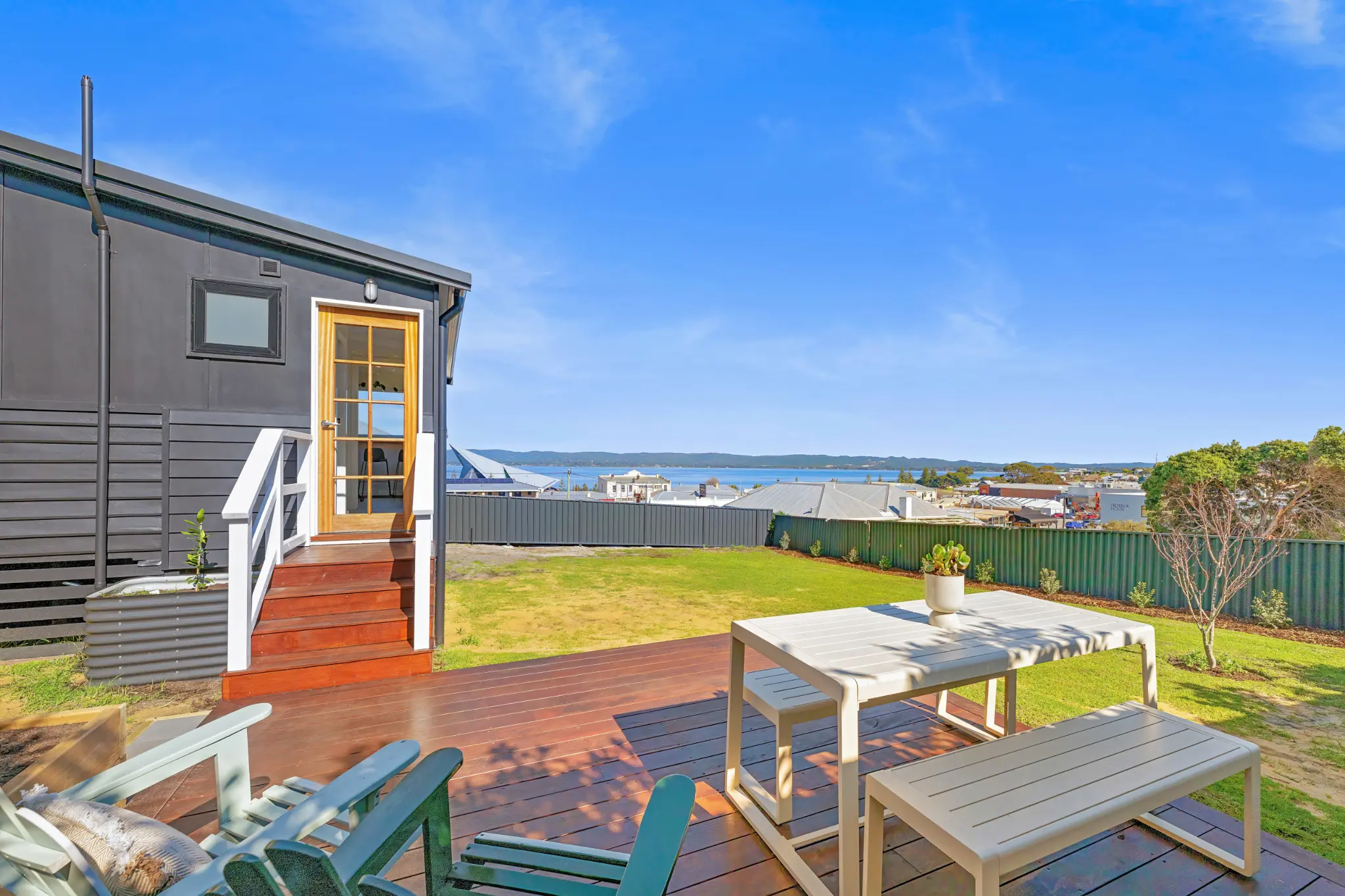 Timber rear deck with outdoor dining table, lounge chairs, fenced backyard and harbour view beyond