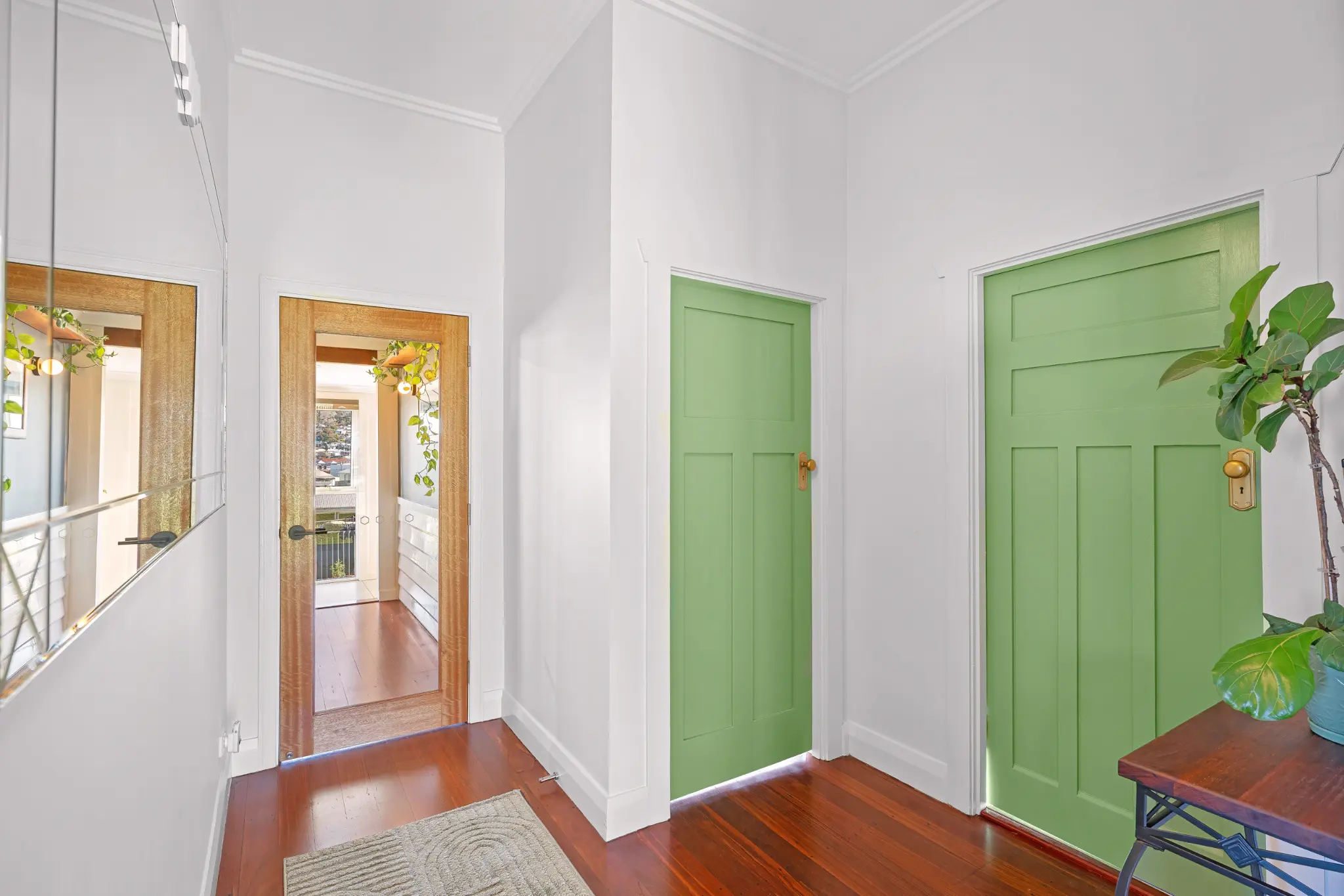 Hallway junction with two green bedroom doors, full-length mirror, fiddle-leaf fig plant and polished jarrah floors
