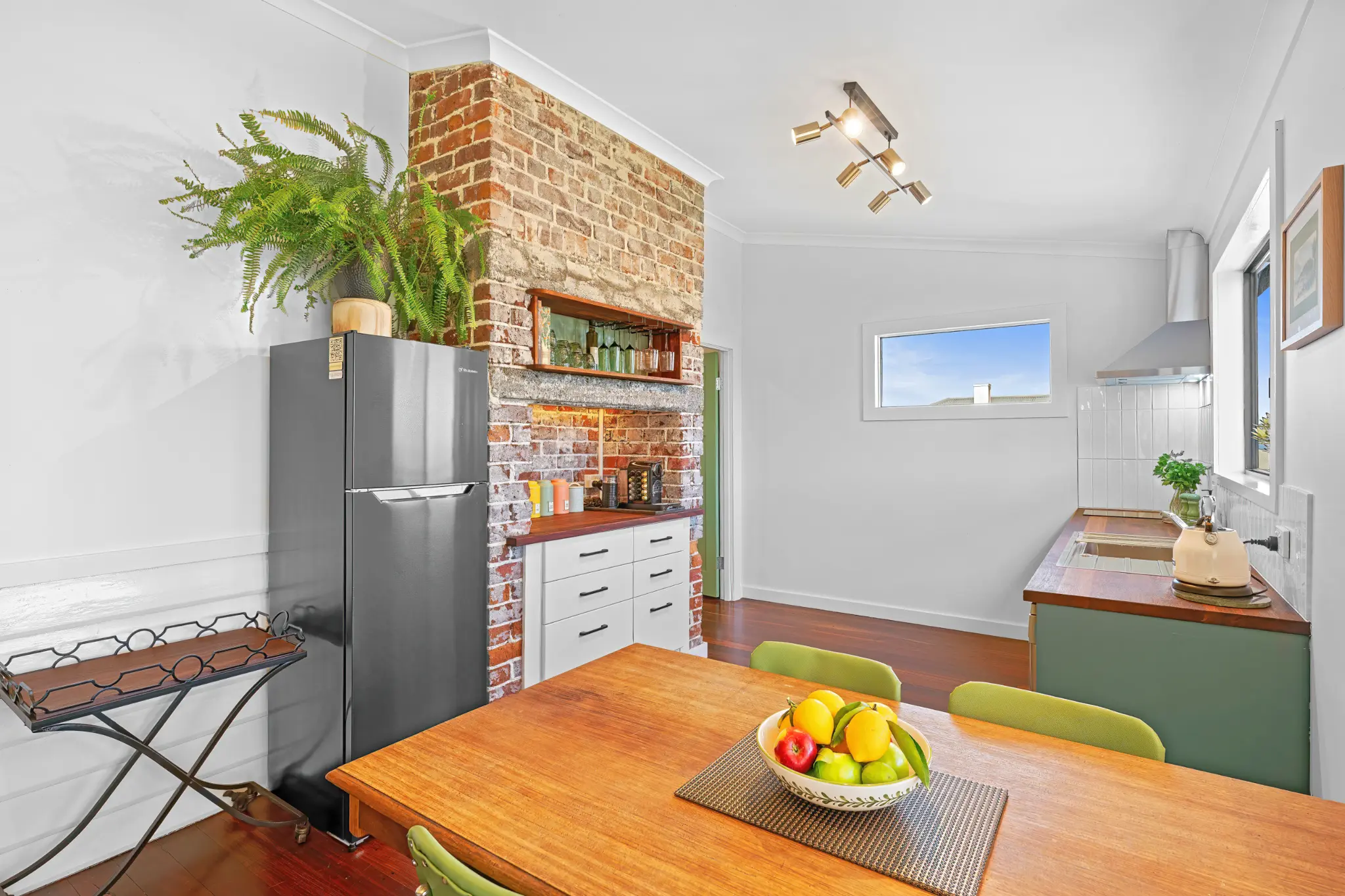 Exposed brick chimney with coffee station, wine glasses on timber shelf and fridge alongside dining table
