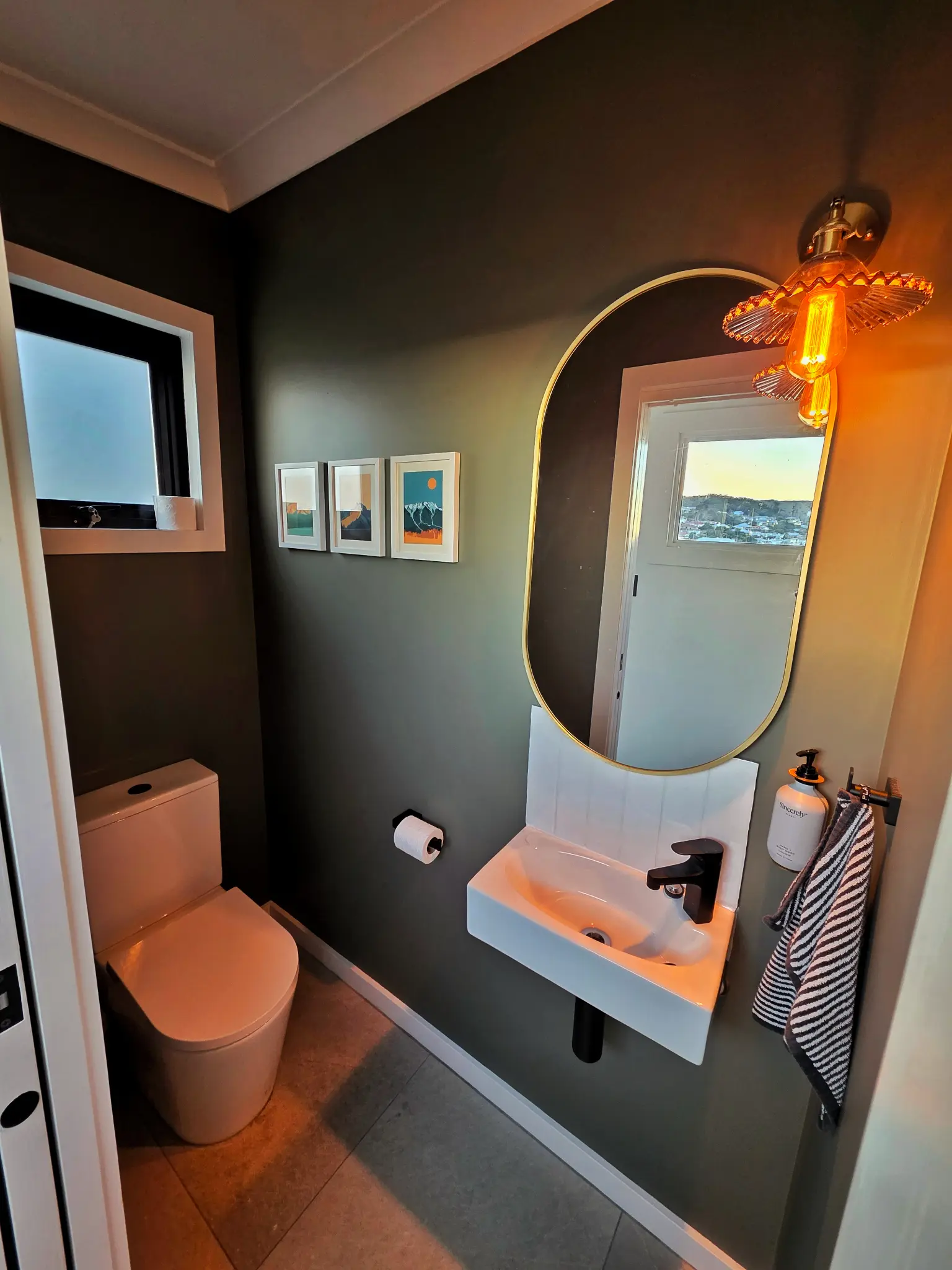 Separate powder room with dark feature wall, gold oval mirror, warm wall sconce and harbour view through window at dusk